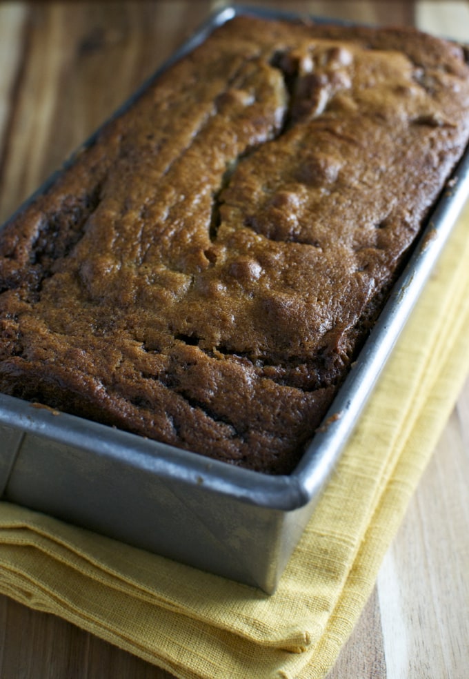 Moist banana bread loaf in a baking pan on a wooden surface with a yellow cloth beneath, perfect for breakfast or a snack.