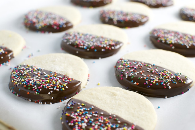 Colorful sprinkles-covered cookies with half dipped in chocolate on a white surface, perfect for sweet treats and party desserts.