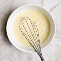 Creamy egg mixture in a white bowl with a metal whisk for baking or cooking recipes.