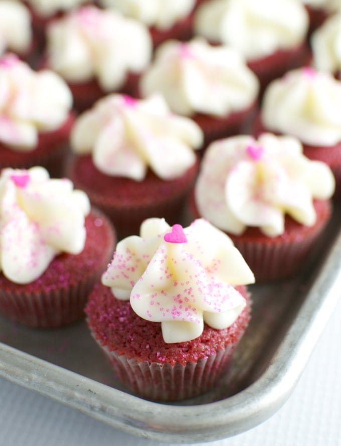 Red velvet cupcakes with white vanilla frosting, pink sprinkles, and heart-shaped decorations on top, perfect for Valentine's Day or special occasions.