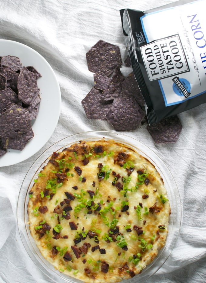 Crispy purple tortilla chips on a white plate with bag of Better Good snack chips, cheesy baked potato pizza with bacon and green onions on a glass plate, white linen background.