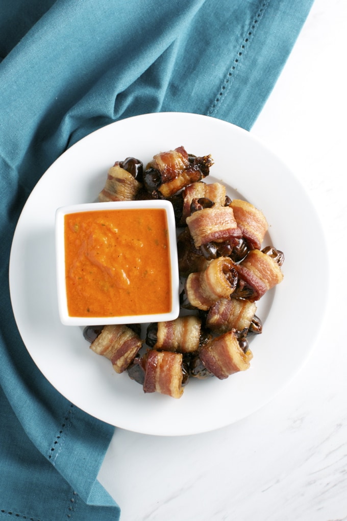 Crispy bacon-wrapped dates with a spicy dipping sauce on a white plate. Perfect appetizer for parties, featuring savory, sweet, and smoky flavors with a decorative presentation.