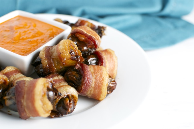 Crispy bacon-wrapped dates stuffed with creamy cheese on a white plate, served with a side of sweet dipping sauce, perfect for appetizers or party snacks.