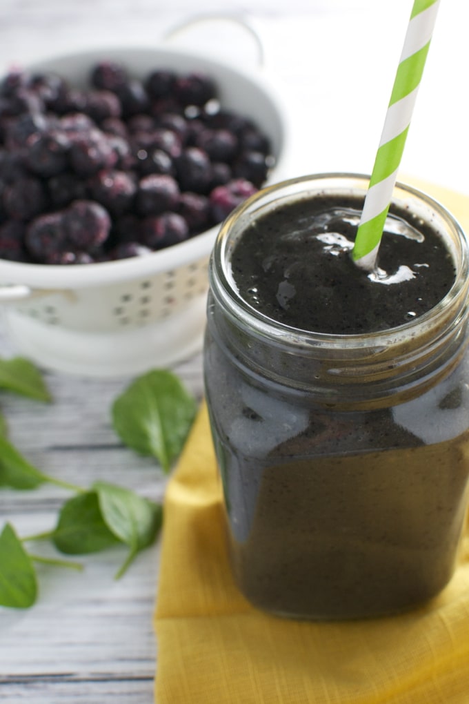 Fresh blueberry smoothie in a mason jar with a striped straw, served on a yellow napkin, with a bowl of whole blueberries and green leaves in the background. Perfect for healthy summer drinks.