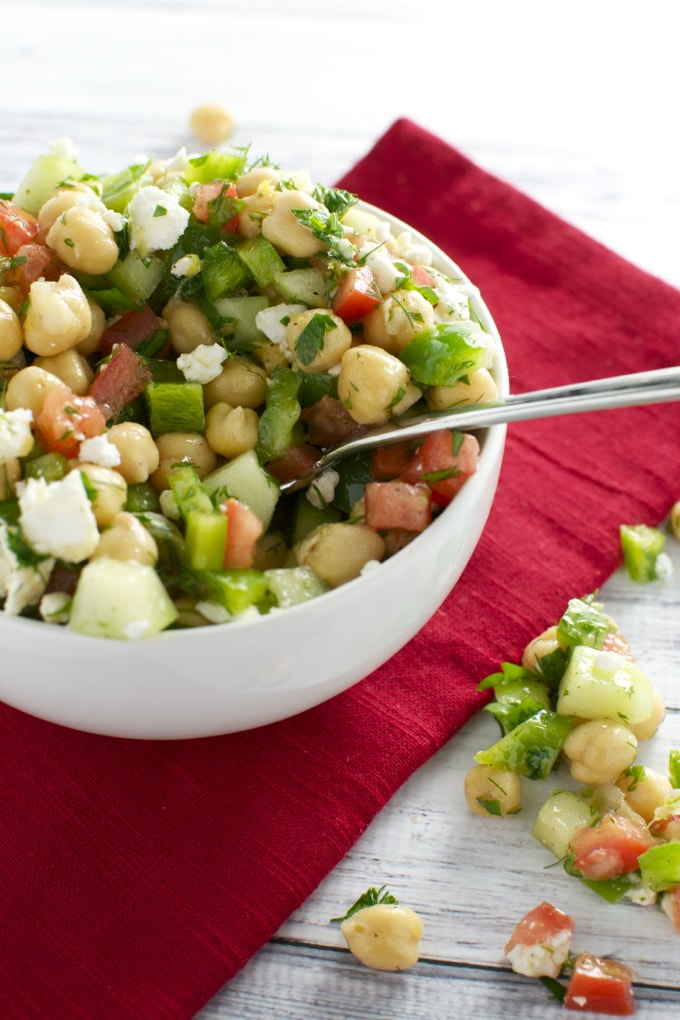 Fresh chickpea salad with diced cucumbers, tomatoes, and parsley in a white bowl, served with a silver spoon on a red cloth napkin, ideal for healthy eating and summer recipes.