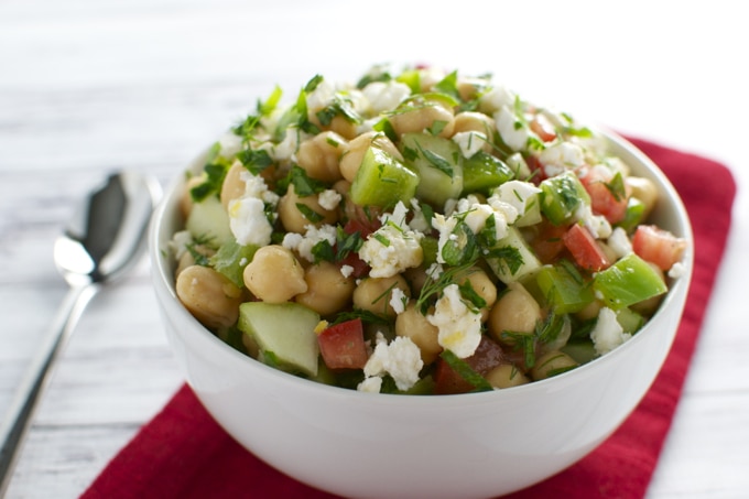 Refreshing chickpea salad with cucumber, tomato, feta cheese, and fresh herbs in a white bowl on a rustic wooden table. Perfect healthy meal or side dish from Stuck On Sweet.