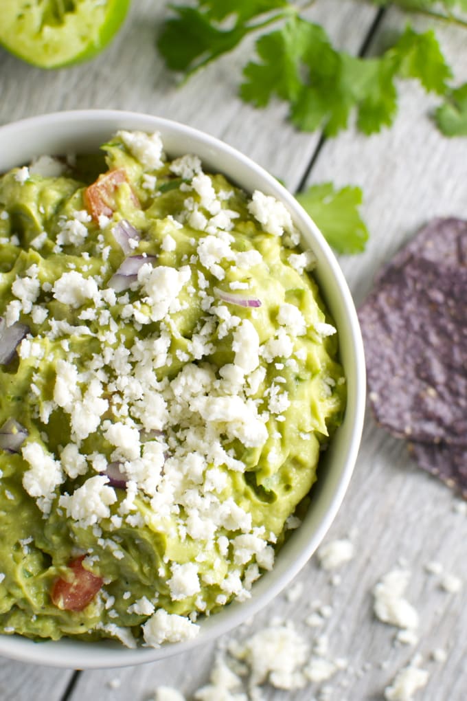 Creamy avocado guacamole with crumbled queso fresco, fresh tomatoes and red onions, served in a white bowl. Perfect for chips and party snacks, fresh and flavorful dip.
