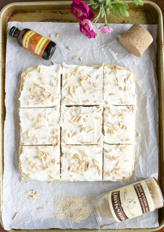 Vanilla layered petit fours with white frosting and toasted almond toppings on baking sheet.