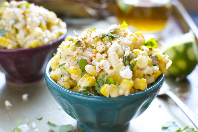 Creamy corn salad in a vibrant blue bowl with fresh herbs, topped with crumbled cheese and spices, served with lime wedges and a glass of drink, perfect for summer meals.