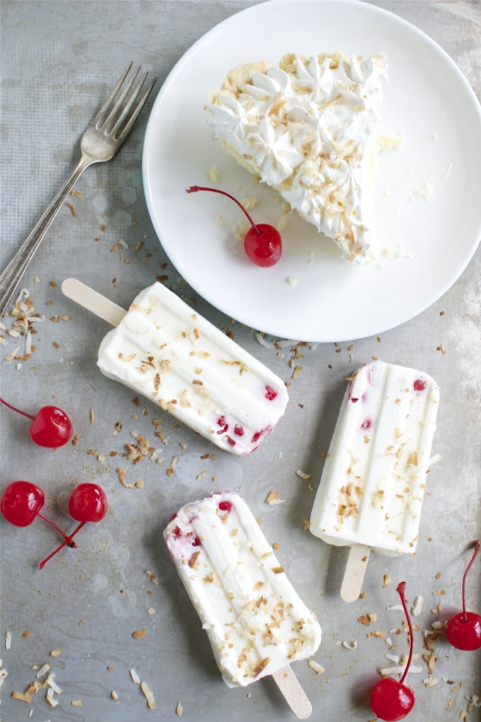 Creamy coconut ice cream bars with cherry toppings and toasted coconut flakes, served on a rustic surface with a slice of coconut cream pie, perfect for summer dessert cravings.