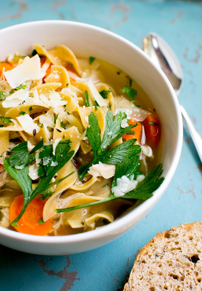 Vegetable pasta soup with fresh herbs, shredded cheese, tomatoes, and a side of multigrain bread in a white bowl.