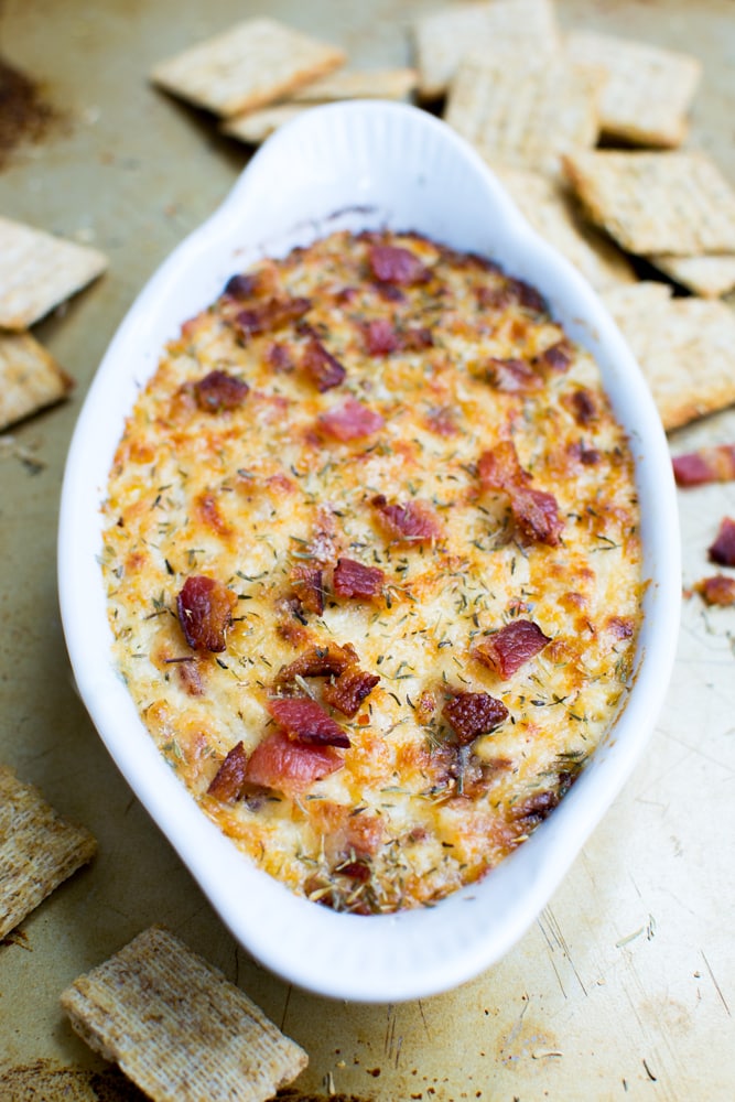 Creamy bacon and onion dip baked to perfection with herbs, served in a white dish, surrounded by crispy crackers on a rustic surface.