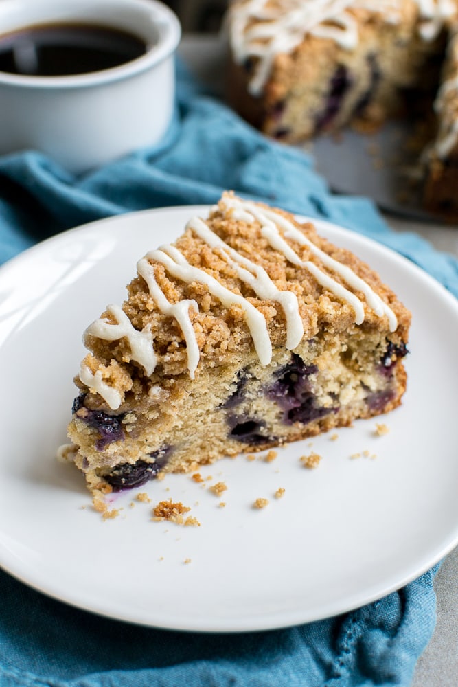 Rich blueberry crumb cake with a sweet streusel topping and white icing drizzle on a white plate, served with a cup of black coffee and a blue napkin.