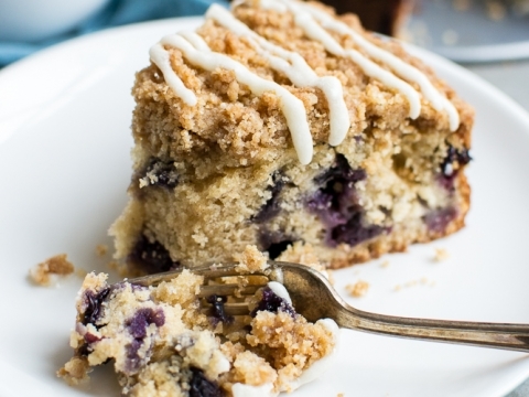Delicious blueberry coffee crumb cake with white icing drizzle on top, served on a white plate with a fork and a cup of coffee in the background.