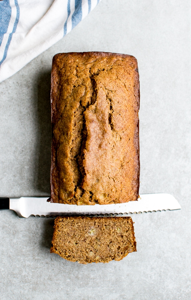 Sweet banana bread loaf on a light gray surface with a serrated knife and a soft striped cloth in the background. Perfect for breakfast or dessert, this moist, homemade banana bread features ripe bananas and warm spices.
