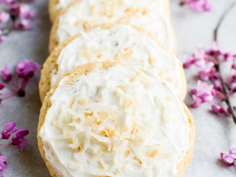 Butter cookies with cream cheese frosting and shredded coconut, topped with toasted coconut flakes, on a white surface with pink flowers.