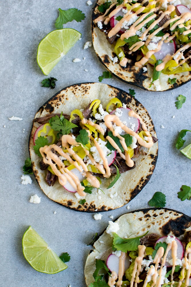 Corn tortilla fish tacos with fresh radishes, cilantro, shredded cheese, and drizzled with spicy sauce on grey surface with lime wedges.