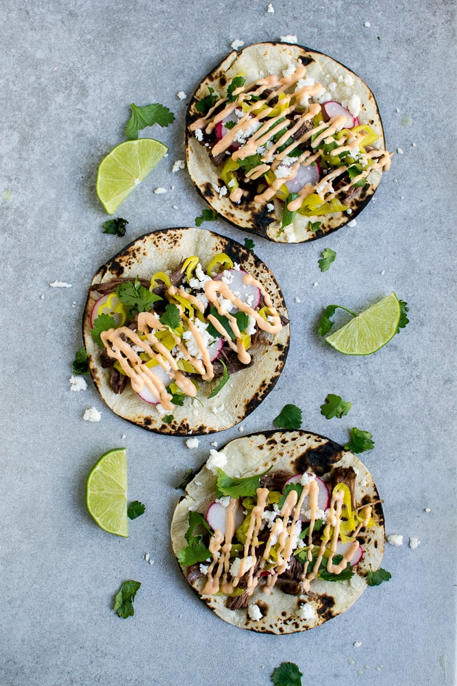 Fresh beef tacos with radishes, cilantro, and spicy sauce on grilled corn tortillas, garnished with lime wedges and crumbled cheese, served on a gray textured background.