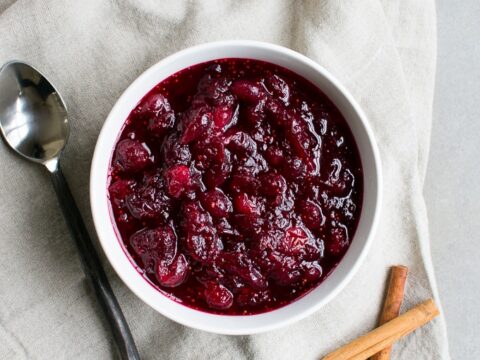 Fresh Homemade Cranberry Sauce in a white bowl with cinnamon sticks on a beige cloth, perfect for desserts or garnishing.