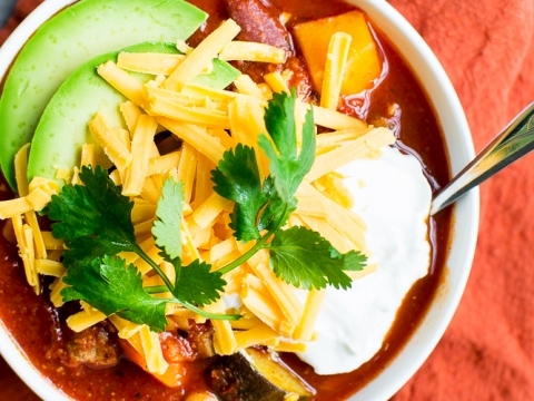 Creamy beef chili bowl topped with shredded cheese, fresh avocado slices, sour cream, and cilantro on an orange napkin. Perfect comfort food recipe from Stuck On Sweet.