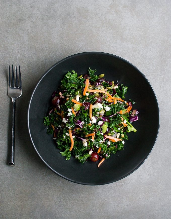Fresh kale salad with shredded carrots, red grapes, feta cheese, and sunflower seeds on a black plate, served with a fork on a gray surface.