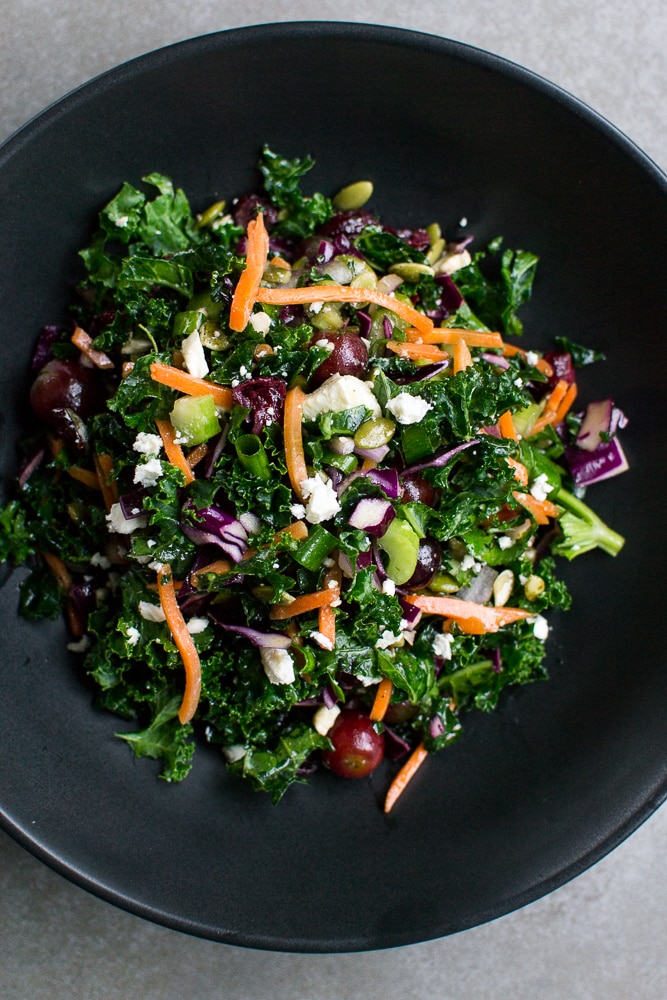 Fresh kale salad with grapes, shredded carrots, celery, and crumbled cheese in a black bowl. Healthy vegetable salad perfect for a nutritious meal or snack.