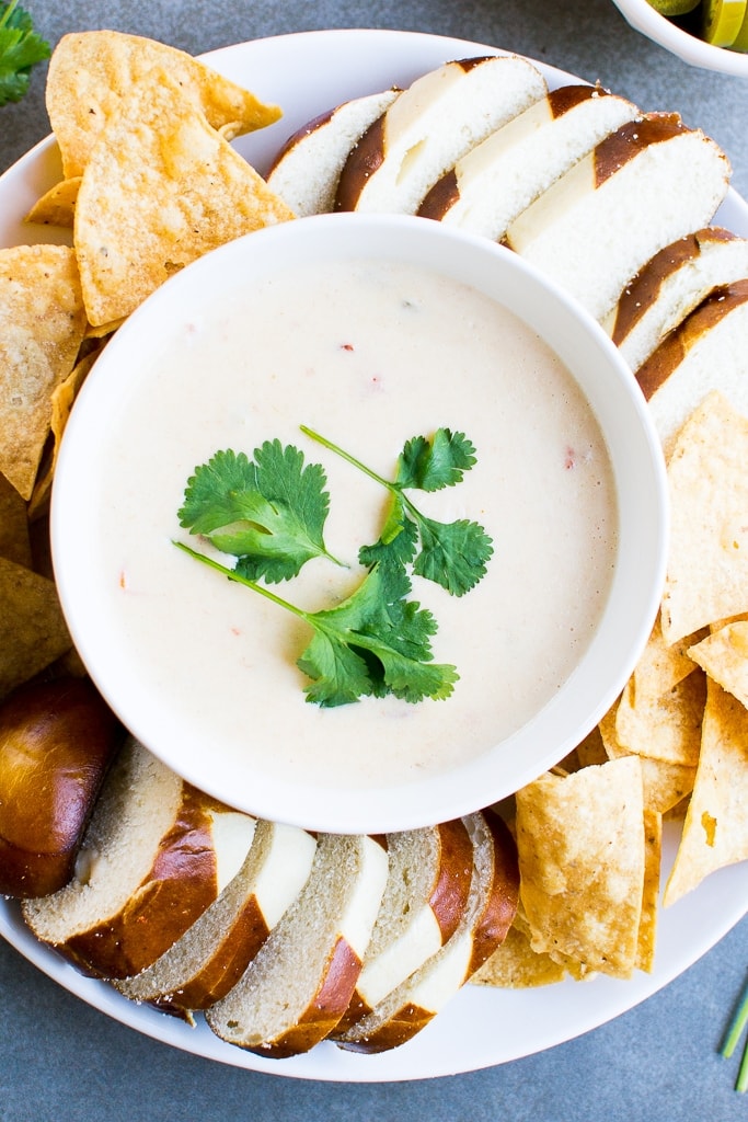 Creamy queso dip garnished with cilantro served with tortilla chips, pretzels, and sliced bread, perfect for snacking or party appetizers.