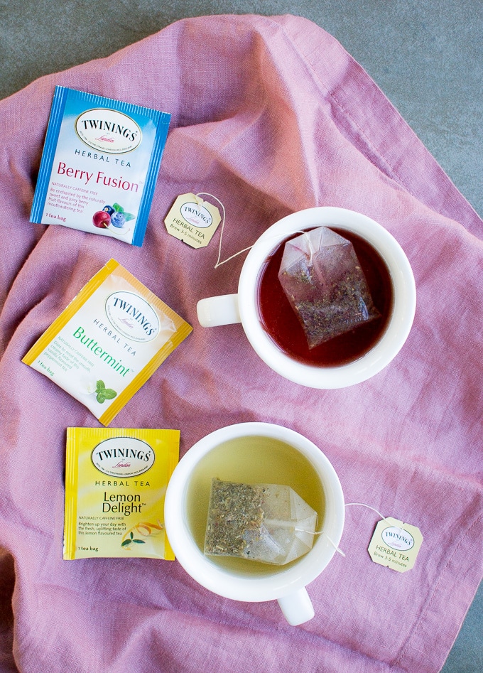 Brightly colored Twinings herbal tea sachets with brewed tea in white mugs on pink fabric, promoting delicious and healthy tea options for wellness and relaxation.