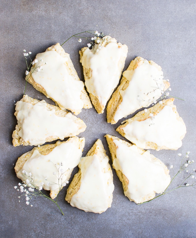 Buttery scones with white glaze and delicate floral garnishes on a gray background. Perfect for breakfast or tea time, these sweet scones from Stuck On Sweet offer a delightful taste of bakery freshness.