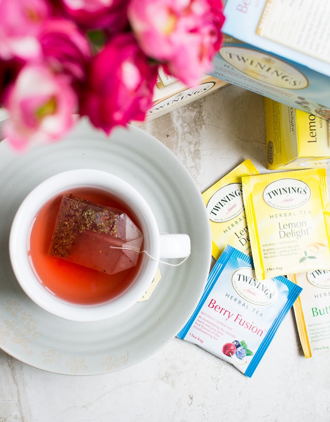 Rich floral scene with a cup of herbal tea on a saucer, surrounded by Twinings tea bags including Lemon Delight and Berry Fusion, showcasing a cozy tea-drinking moment.
