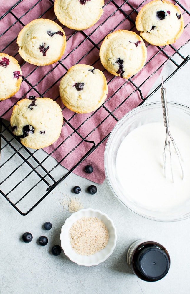 Fresh blueberry muffins cooling on a wire rack with a bowl of vanilla glaze, blueberries, and ingredients for baking, perfect for sweet breakfast or dessert.