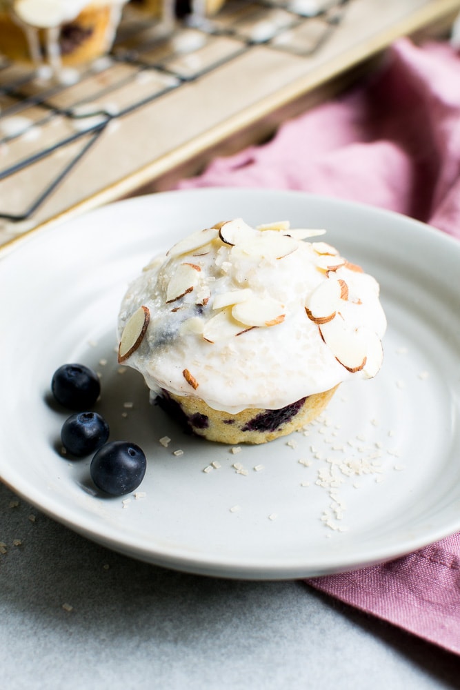 Sweet blueberry muffin with creamy frosting and sliced almonds served on a white plate with fresh blueberries, perfect for breakfast or dessert.