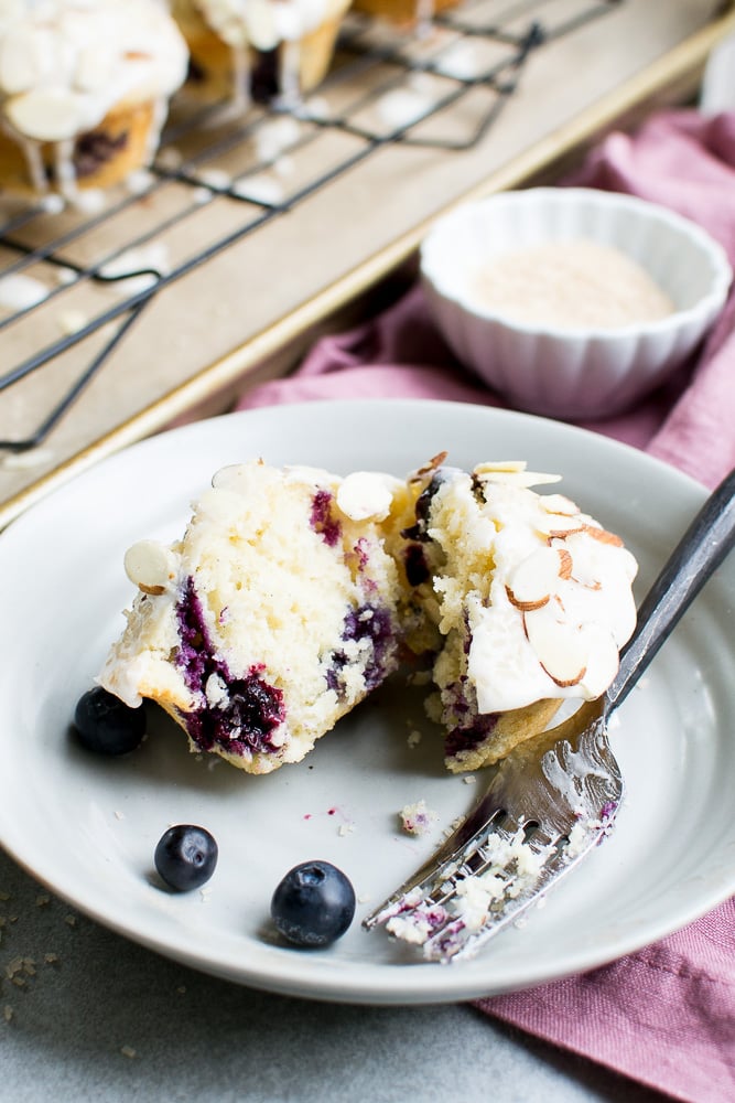 Fresh blueberry lemon cupcake with creamy frosting and sliced almonds on top, served on a white plate with blueberries and a fork, featuring a pink linen napkin, sweet bakery atmosphere, Stuck On Sweet.