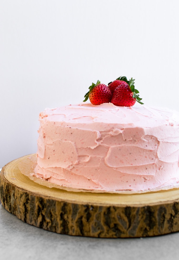 Fresh strawberry cake with pink frosting and whole strawberries on top, displayed on a rustic wooden serving board, perfect for birthdays and special occasions.
