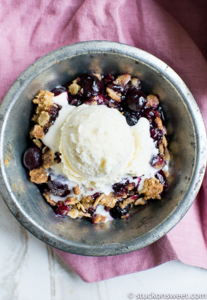 Sweet cherry blueberry crumble dessert with vanilla ice cream in a rustic metal bowl. Perfect for summer, featuring a warm fruit topping and creamy ice cream.