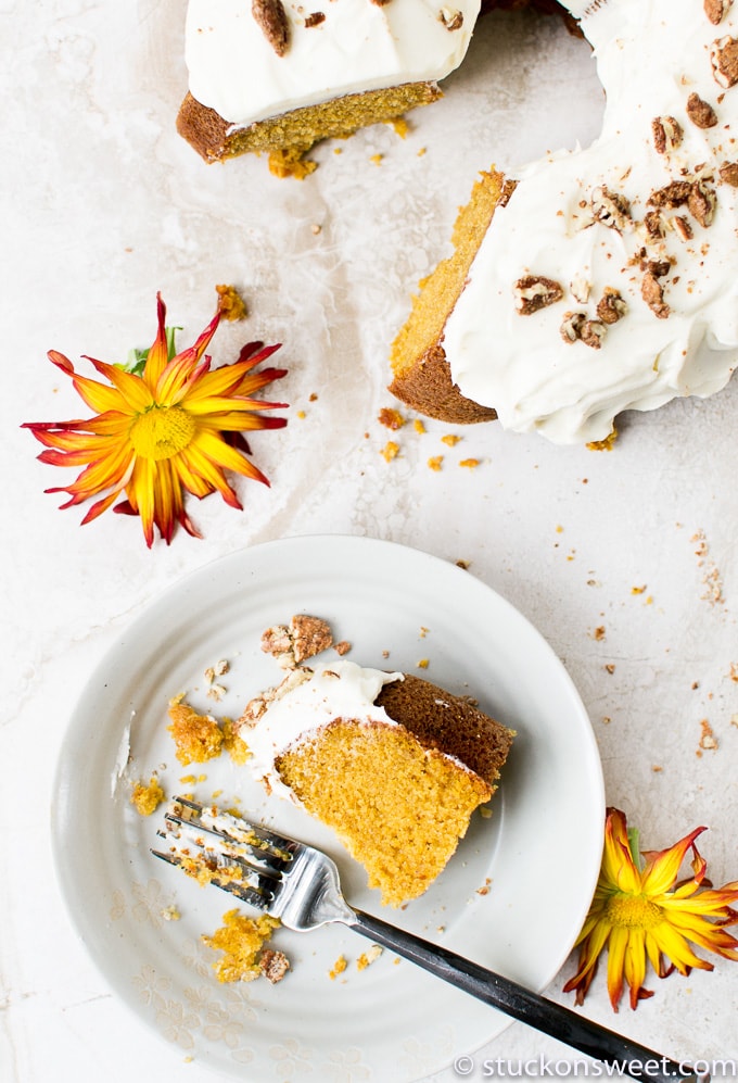 Moist homemade spiced pumpkin bundt cake with cream cheese frosting and chocolate sprinkles, served on a white plate with a fork, topped with a vibrant yellow daisy flower.