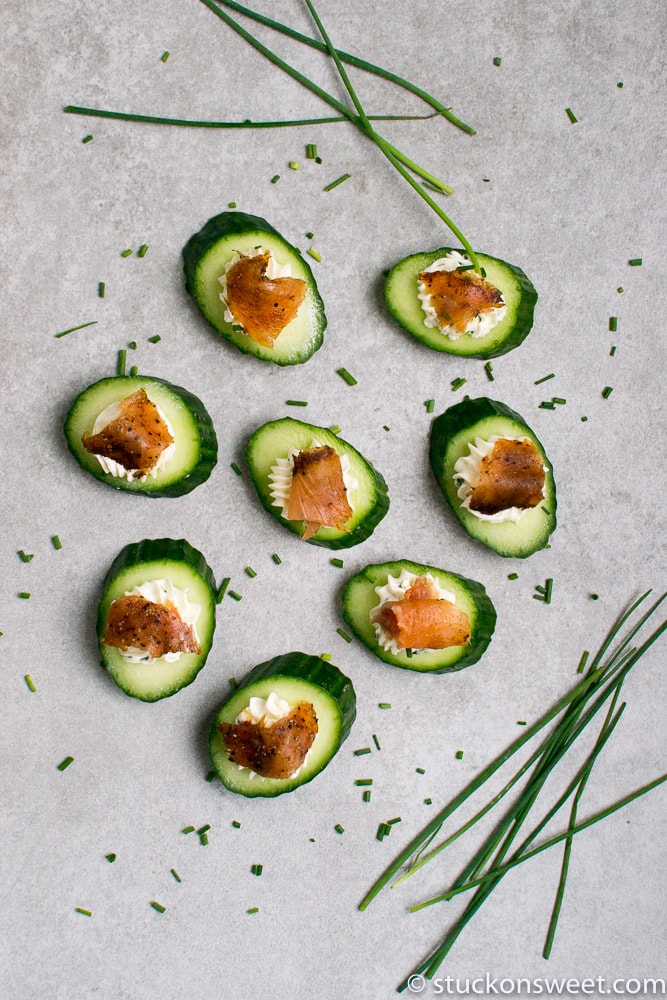 Cucumber bites topped with cream cheese and smoked salmon on a light gray surface, garnished with chives and sprinkled with chopped chives for a fresh, elegant appetizer.