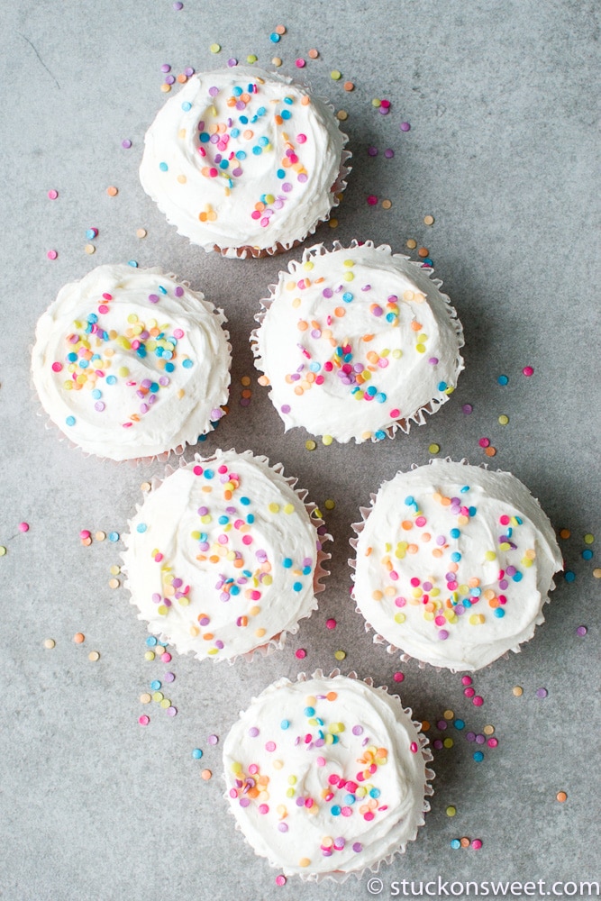 Colorful sprinkle cupcakes with white frosting and scattered sprinkles on gray background.