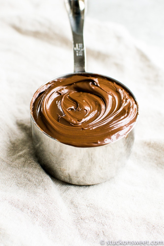 Creamy melted chocolate for baking or desserts, in a metal measuring cup on a neutral fabric background. Perfect for creating rich, delicious homemade chocolates, icings, and confections.