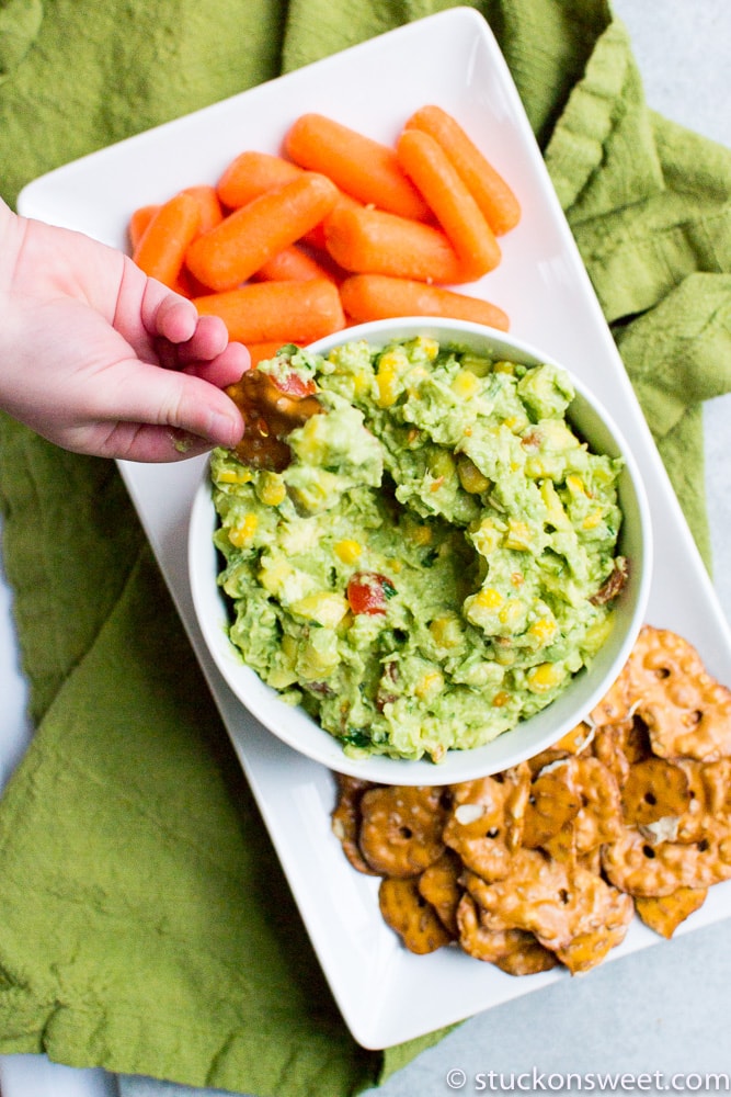 Creamy guacamole with fresh vegetables, orange cheese balls, and sweet glazed pretzels on a white serving platter for party snacks.