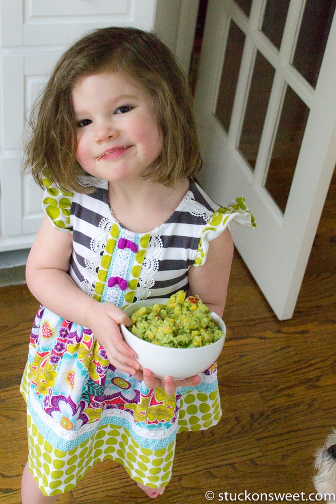 Fresh homemade guacamole served in a white bowl, with a young girl in colorful dress holding it, inside a cozy kitchen – perfect for family fun and healthy snack ideas.