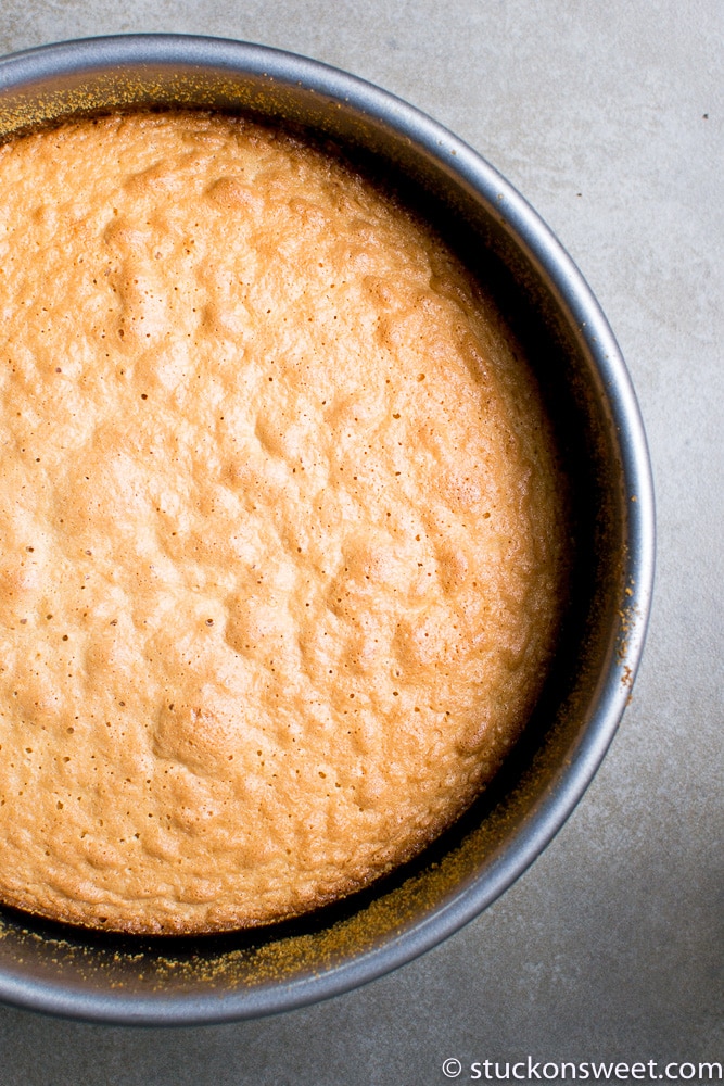 Fluffy, golden baked cake batter in a round metal baking pan with a light gray textured background.