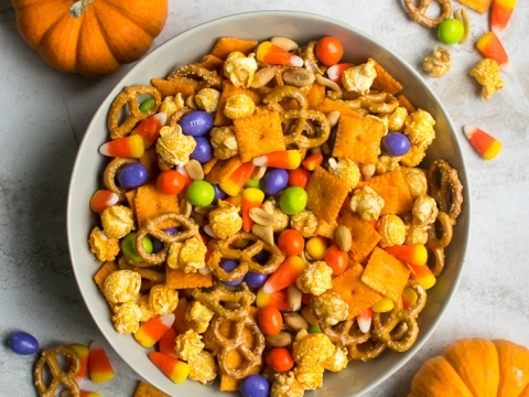 Candy corn Halloween snack mix with pumpkin decor for autumn and fall celebrations, featuring pretzels, popcorn, and colorful candies on a white plate.