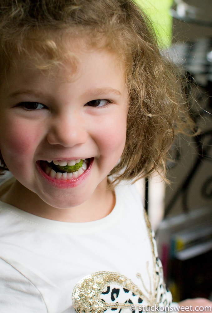 Vibrant close-up of a smiling young girl with curly hair enjoying a delicious green apple slice in her mouth, showcasing childhood joy and healthy snack choices at Stuck On Sweet.