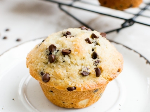 Chocolate chip muffin with golden crust on a white plate, with more muffins in the background cooling on a wire rack. Perfect for breakfast or snack, classic homemade muffin recipe.