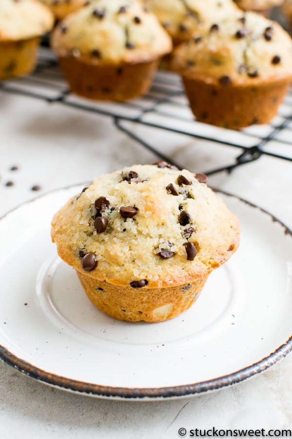 Chocolate chip muffin with golden crust on a white plate, with more muffins in the background cooling on a wire rack. Perfect for breakfast or snack, classic homemade muffin recipe.