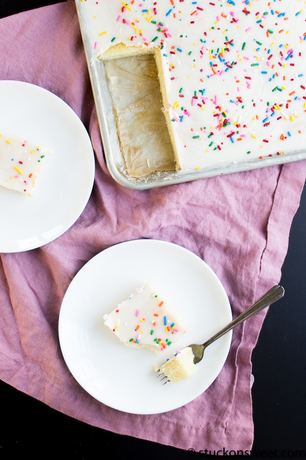 Bright birthday cake with colorful sprinkles and smooth white frosting, served on white plates with a fork, set on a pink linen cloth for a festive celebration.