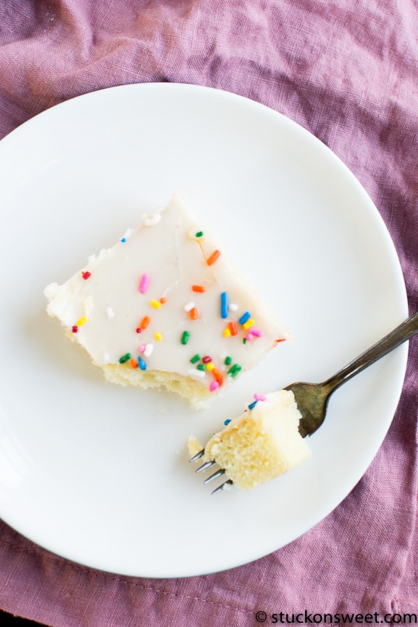 Butter vanilla cake slice with white frosting and colorful sprinkles on a white plate, with a small fork, pink cloth background.