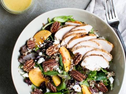 Fresh grilled chicken salad with peaches, pecans, and feta cheese on a white plate, served with honey vinaigrette on the side, perfect for a healthy meal or summer lunch.