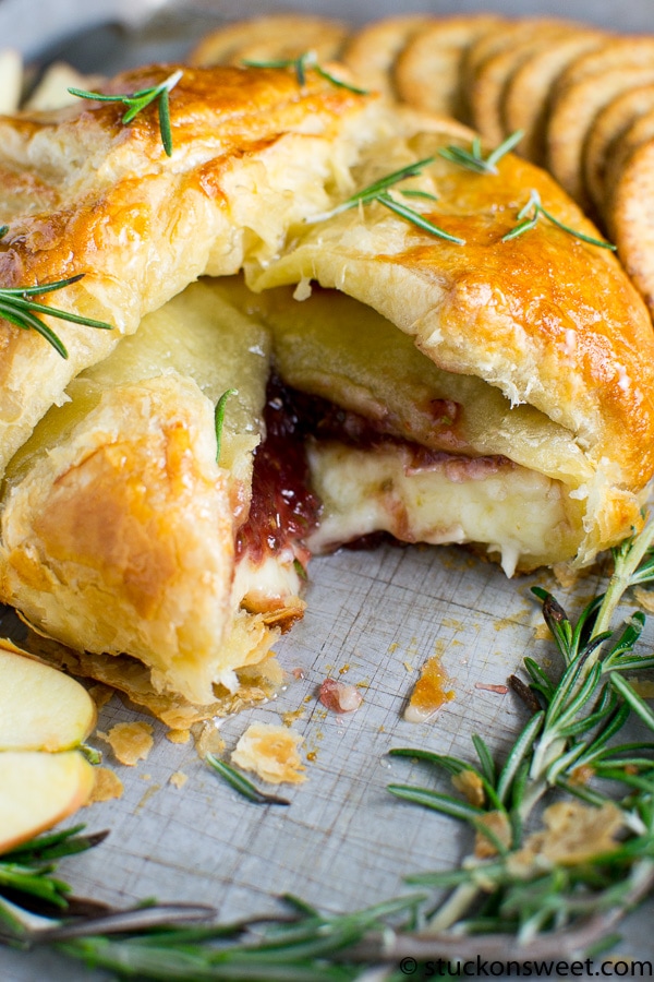 Golden puff pastry Baked Brie with preserves pie with melted cheese and fruit filling, garnished with fresh rosemary, served with sliced apples and cookies, perfect for dessert or snack.